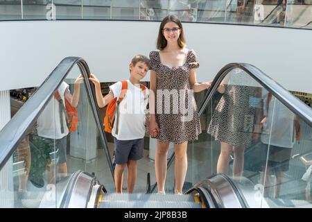 Une famille heureuse avec des sacs en papier est debout sur un escalier roulant dans un centre commercial et discute de leurs achats, réflexions. Maman et fils avec une école b Banque D'Images