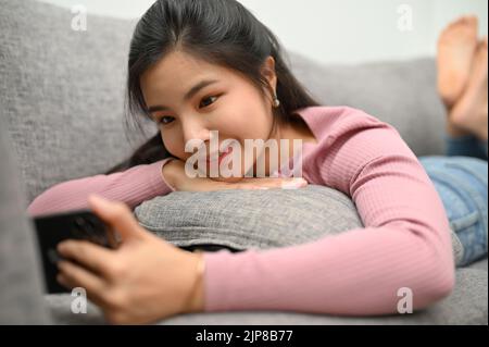 Jolie charmante jeune femme asiatique se détend sur son confortable canapé a dans le salon et à l'aide d'un smartphone, en vérifiant ses médias sociaux. Banque D'Images