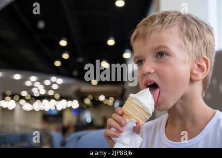 Portrait d'un petit garçon drôle et affamé qui mange de la glace froide délicieuse dans une tasse de gaufres assis dans un café, fond sombre, bokeh. Un enfant aime la glace Banque D'Images