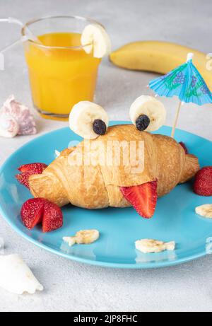 Nourriture amusante pour les enfants - croissant en forme de crabe avec fraise et banane sur une assiette bleue et un verre de jus d'orange sur fond gris, format vertical Banque D'Images