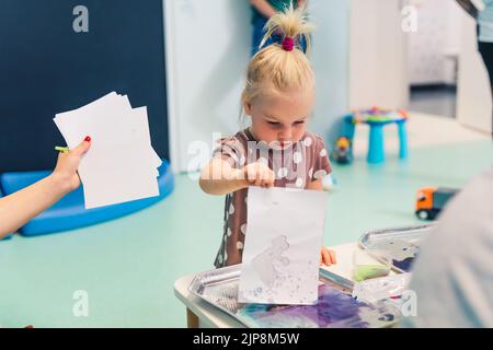 Petite caucasienne mignonne de peinture en utilisant des peintures non toxiques avec un enseignant dans la salle de classe à l'école maternelle. Photo de haute qualité Banque D'Images