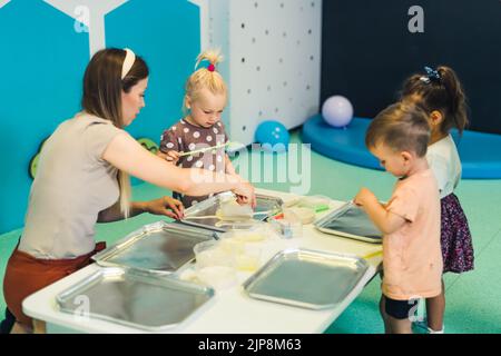 Jeu sensoriel à l'école maternelle multi-culturelle. Les tout-petits avec leur professeur jouant avec des pailles rayées et la peinture au lait, en utilisant la coloration de nourriture, le lait, le papier aquarelle, et les plateaux. Activité créative pour les enfants pour le développement du cerveau. Peinture au doigt. Photo de haute qualité Banque D'Images