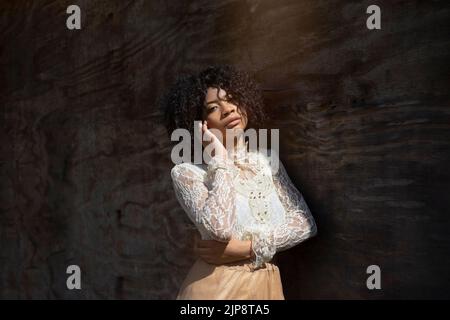 Jeune femme confiante modèle ethnique avec cheveux bouclés afro en dentelle blanche blouse touchant le visage et regardant l'appareil photo tout en se tenant près du mur en plein soleil Banque D'Images