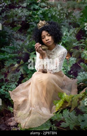 Ensemble complet de jeune afro-américaine modèle pacifique avec les yeux fermés dans une tenue romantique et la couronne assis au milieu de plantes vertes luxuriantes avec n Banque D'Images