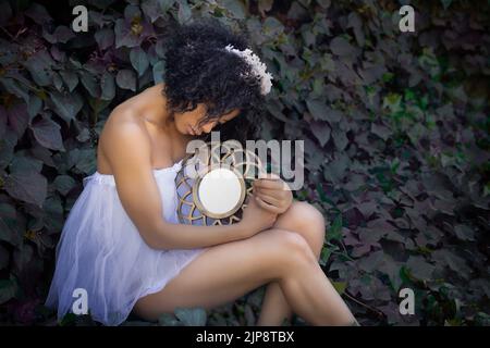 Vue latérale d'une jeune afro-américaine rêveuse avec des cheveux maurmés dans une robe blanche romantique et un accessoire tête tenant un miroir vintage pendant qu'elle est assise Banque D'Images