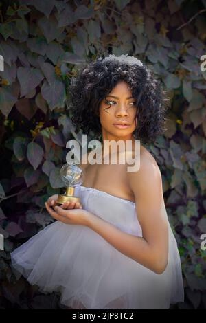 Vue latérale de la jeune femme afro-américaine féminine avec des cheveux noirs bouclés en robe romantique avec sphère magique dans les mains regardant loin en se tenant agai Banque D'Images