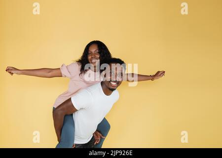 Jeune couple souriant et gai, femme avec les mains de côté imitant le vol assis sur le dos d'un petit ami, debout au studio, isolée sur fond jaune, regardant la caméra. Banque D'Images