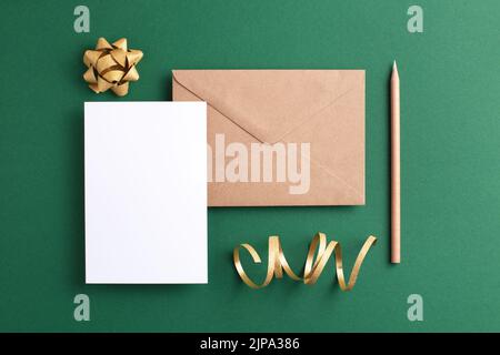 Carte vide, enveloppe kraft, crayon de bois et décorations dorées sur fond vert. Maquette de vacances et d'invitation. Banque D'Images