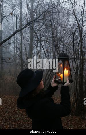 Une jeune femme marche à travers la forêt sombre, elle tient une lanterne en fer. Paysage brumeux en arrière-plan. Banque D'Images