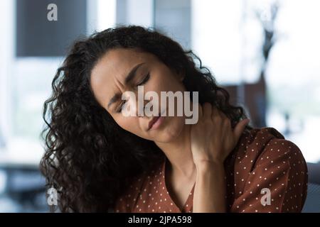 Photo rapprochée d'une femme aux cheveux bouclés à la maison près de la fenêtre avec une douleur au cou sévère, femme d'affaires mulatto massant son cou avec les yeux fermés Banque D'Images