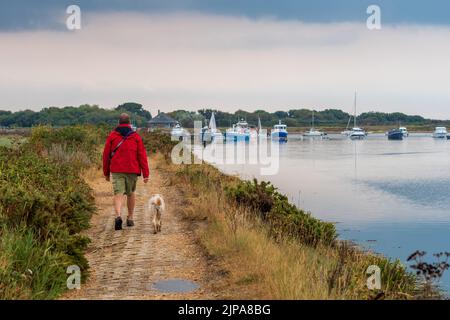 Keyhaven, Milford-on-Sea, Hampshire, Royaume-Uni, 16th août 2022, Météo. Des averses lourdes accompagnent les nuages sombres comme la pluie nécessaire tombe sur sec, parché sud de l'Angleterre. Un homme sous un imperméable rouge marche son chien le long du front de mer. Crédit : Paul Biggins/Alamy Live News Banque D'Images