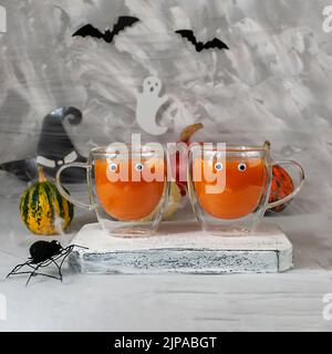 Deux tasses en verre avec jus de citrouille sur un fond de citrouilles d'Halloween sur un mur gris avec des fantômes, des chauves-souris, des araignées, des chapeaux de sorcière, des citrouilles.Conceptuel Banque D'Images