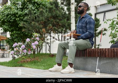 Portrait complet d'un homme aveugle portant des lunettes de soleil et livre de lecture en braille à l'extérieur, espace de copie Banque D'Images