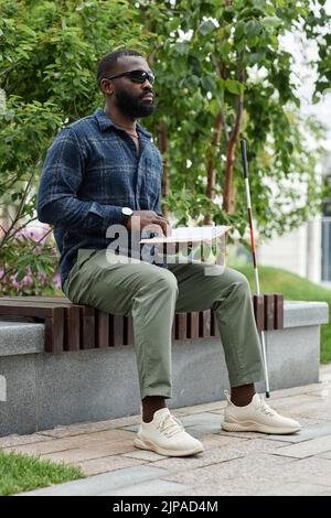 Portrait vertical complet d'un aveugle portant des lunettes de soleil et livre de lecture en braille à l'extérieur Banque D'Images