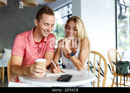 Des étudiants heureux assis dans la table de café de l'université et faisant des devoirs ou de la rédaction de plans. Une jeune femme et un homme passent du temps ensemble pendant le frein du café, en prenant des notes. Banque D'Images