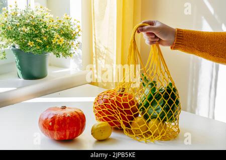 Main femelle tenant le filet de shopping avec des légumes - citrouilles et salade verte - à la cuisine. Couleurs jaune et orange. Concept zéro déchet Banque D'Images