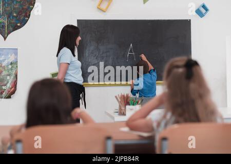 École primaire. L'enseignante aidant l'enfant étudiant tout en écrivant la réponse sur le tableau de surveillance. Banque D'Images