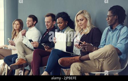 Sur le wifi pendant l'attente. Un groupe d'hommes et de femmes d'affaires utilisant différents appareils sans fil en attendant une interview. Banque D'Images