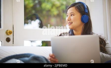 Elle a tout couvert pour le week-end. C'est une jeune femme très attirante qui porte des écouteurs tout en utilisant un ordinateur portable à la maison. Banque D'Images