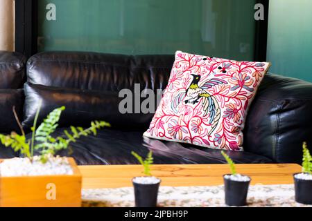 L'oreiller rouge à motifs paon est sur un canapé noir. En face de lui était une petite plante en pot sur une table en bois marron. Banque D'Images