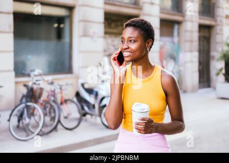 Belle femme africaine marchant dans les rues et souriant en parlant par téléphone. Elle porte une tenue décontractée d'été et une coupe de cheveux tendance Banque D'Images