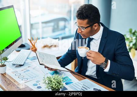 Il a une assiette complète aujourd'hui. Un jeune homme d'affaires concentré assis à son bureau tout en buvant du café et en lisant son journal dans le bureau. Banque D'Images