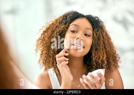 Maintenant pour les touches de finition. Une jeune femme confiante appliquant de la crème de peau sur son visage tout en regardant dans le miroir dans la salle de bains à la maison. Banque D'Images