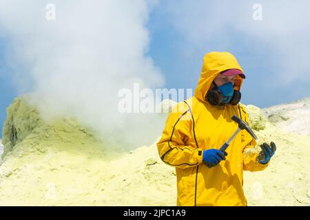 femme volcanologue à l'arrière-plan d'un fumarale fumeur examine un échantillon d'un minéral de soufre avec un marteau géologique Banque D'Images