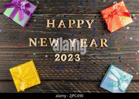 Boîtes-cadeaux multicolores avec noeuds noués. Nouvel an présente sur une table en bois avec des étoiles confetti éparpillées. Texte bonne année 2023. Accueil festif ca Banque D'Images