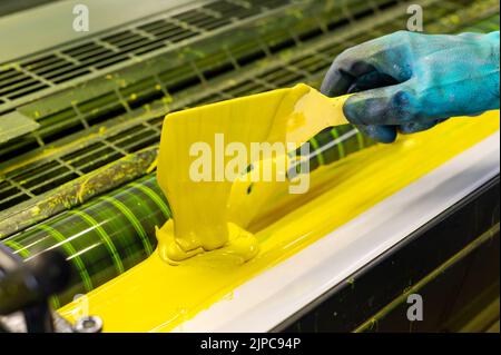 Par dessus l'employé anonyme en caoutchouc bleu gant à l'aide d'une spatule pour enduire la peinture jaune sur les rouleaux de l'imprimante offset industrielle Banque D'Images