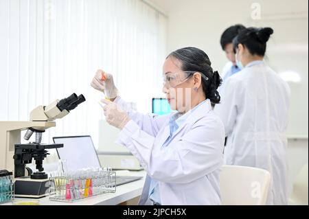 Scientifique médical ou chimiste féminin asiatique d'âge professionnel dans des lunettes et une robe ajustant un échantillon médical dans un tube à essai en laboratoire. Banque D'Images