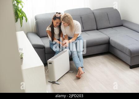 Chauffage électrique à la maison. Une jeune famille réchauffe ses mains gelées près d'un radiateur de chauffage Banque D'Images