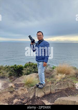 Prise de vue verticale d'un homme d'âge moyen dans une veste bleue avec un appareil photo posé sur une falaise Banque D'Images