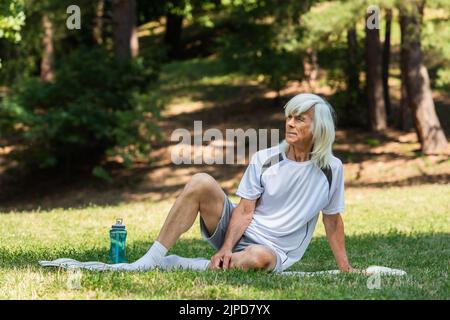 pleine longueur d'homme à la retraite dans des vêtements de sport assis sur un tapis de fitness dans le parc vert Banque D'Images