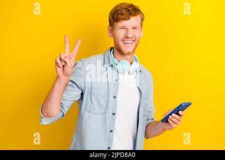 Photo de gai positif garçon homme tendance jeans ensemble montrer v symbole bras tenir téléphone profiter de la liste de lecture isolée sur fond jaune couleur Banque D'Images