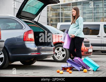 Une femme caucasienne avec des sacs en papier donne naissance prématurée à l'enfant au parking automatique dans le centre-ville. Jeune femme avec l'abdomen ventre sensation de douleur pendant l'eau brisée après le shopping pendant le temps d'attente. Banque D'Images