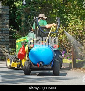 Jardinier assis en hauteur sur un mini-tracteur John Deere tractant une mini-remorque de camion-citerne à eau pompée bleue pulvérisant des fleurs dans l'hôtel Country Estate Angleterre Royaume-Uni Banque D'Images