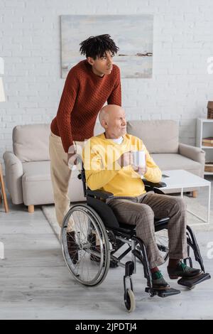 Homme afro-américain debout près du grand-père tenant une tasse en fauteuil roulant à la maison Banque D'Images