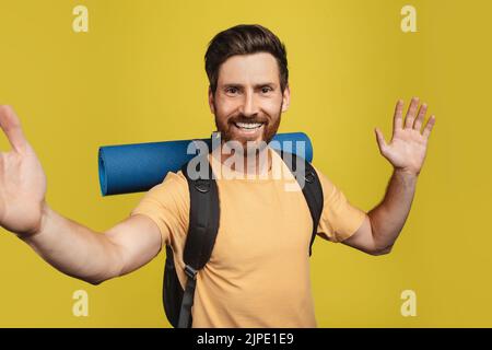 Joyeux homme d'âge moyen avec des engins de camping qui emmenaient le selfie et qui agite vers l'appareil photo de smartphone sur fond jaune de studio Banque D'Images