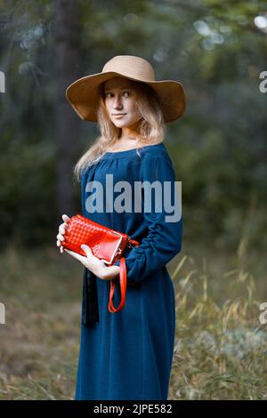 Une jeune fille de cheveux longs dans un chapeau de paille et avec un sac rouge dans sa main marche dans le parc. Arrière-plan flou de la nature. Banque D'Images