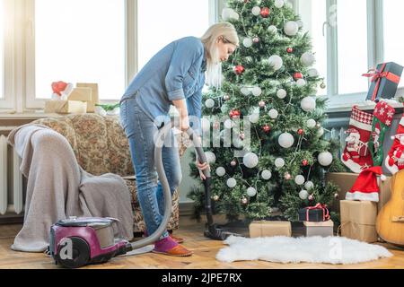 Une jeune femme qui nettoie avec un aspirateur, passe l'aspirateur sous les aiguilles de Noël avec des ornements du nouvel an sur un parquet en bois dur. Banque D'Images