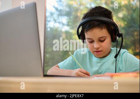 Gros plan d'un écolier concentré dans un casque audio prenant des notes pendant la leçon en ligne avec un professeur de Web à distance Banque D'Images