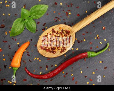 Vue de dessus des flocons de piment et des graines séchés dans une cuillère de cuisson en bois avec des piments rouges et orange sur un panneau d'ardoise noir Banque D'Images