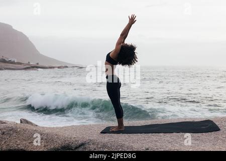 Vue latérale d'une femme mince levant les mains dans le yoga pose debout contre un océan le soir Banque D'Images