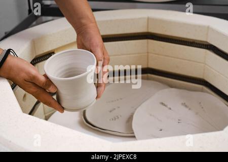 Gros plan des mains mettant vase dans le four, four pour rôtir des produits faits à la main en argile non cuite, céramiques non finies et ustensile. Poterie moderne Banque D'Images