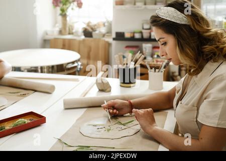 Femme décorant avec des fleurs roulé argile, faisant plaque en céramique en studio avec motif floral. Travail créatif fait main. Atelier de poterie pour adultes. Banque D'Images