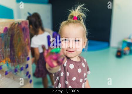 Peinture de film à l'école maternelle. Bonne fillette blonde souriante montrant sa paume colorée peinte. Cours d'art avec peinture au doigt pour les enfants compétences sensorielles, créativité et développement de l'imagination. Photo de haute qualité Banque D'Images