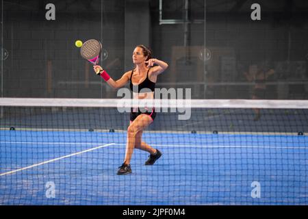 Femme jouant de padel dans un terrain de padel d'herbe bleue intérieur - jeune femme sportive joueur de padel frappant le ballon avec une raquette Banque D'Images