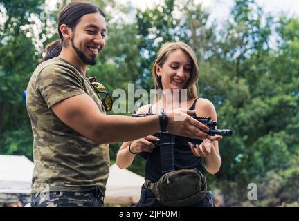 Homme barbu dans un t-shirt camouflage montrant une arme de sous-machine à la cliente femelle. Achat et vente d'armes à feu. Prise de vue horizontale en extérieur. Photo de haute qualité Banque D'Images