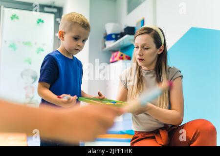 École maternelle. Un tout-petit garçon caucasien et son professeur préparent des peintures et des pinceaux pour un film adhésif peint sur un film adhésif enveloppé autour du support d'étagères comme un chevalet. Activités artistiques amusantes pour les enfants compétences sensorielles, créativité et développement de l'imagination . Photo de haute qualité Banque D'Images